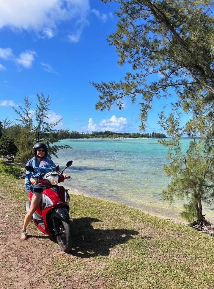 woman on a motor scooter standing on the grass right next to the shallow crystal clear ocean water in Mauritius
