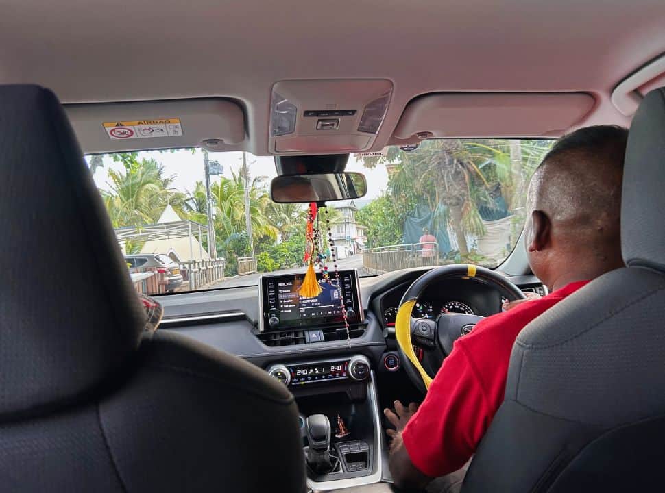 picture taken inside a taxi, with the driver behind the steering wheel driving through a town in Mauritius