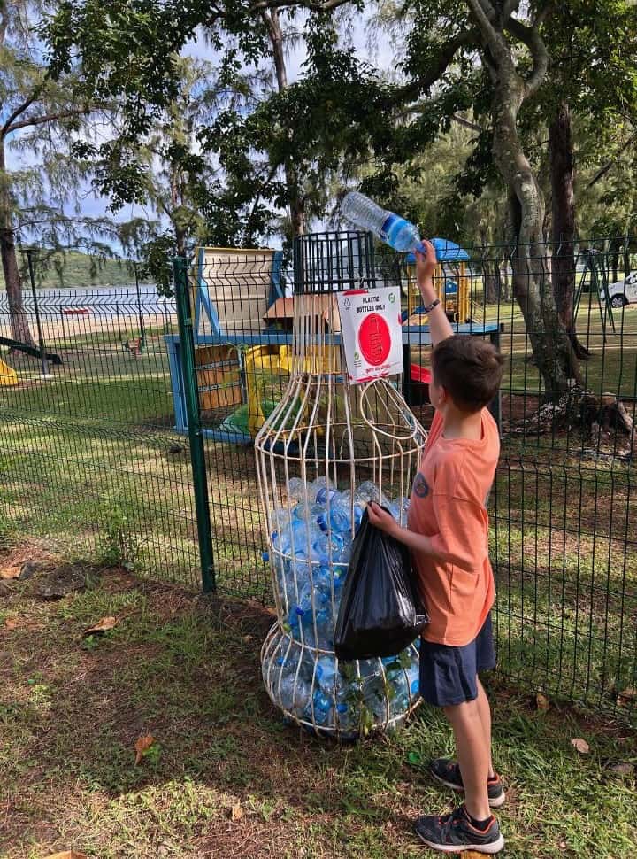 boy throwing away empty plastic bottles in a designated container for plastic bottles only, located near a playground and beach in Le Morne Brabant Mauritius