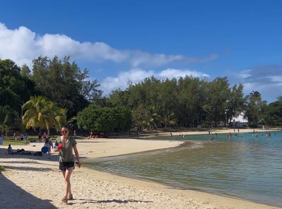 woman walking along the water edge at Blue Bay beach Mauritius, in the back people are swimming in the water, tall trees grow along this beach in Mauritius