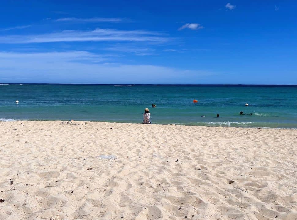woman sitting at the edge of the beach near the blue green ocean water, watching children play in the water at Flic en Flac Beach