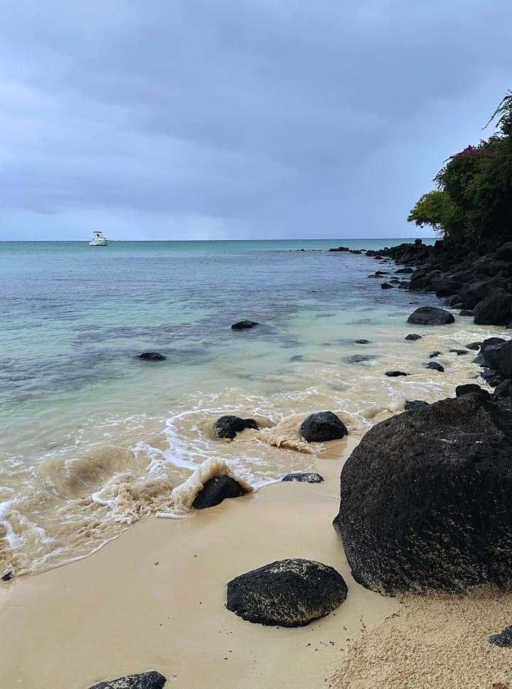 black rocks along the edge of a beach, the small waves crash slightly against them, the rocks in the distance are visible due to the clear water despite the overcast sky in Mauritius