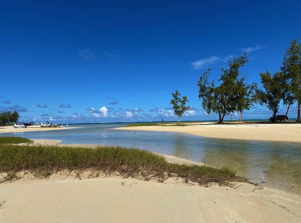 soft sanded beach with a sandbank with a row of trees, calm waters. Cars parked on the beach in Mauritius