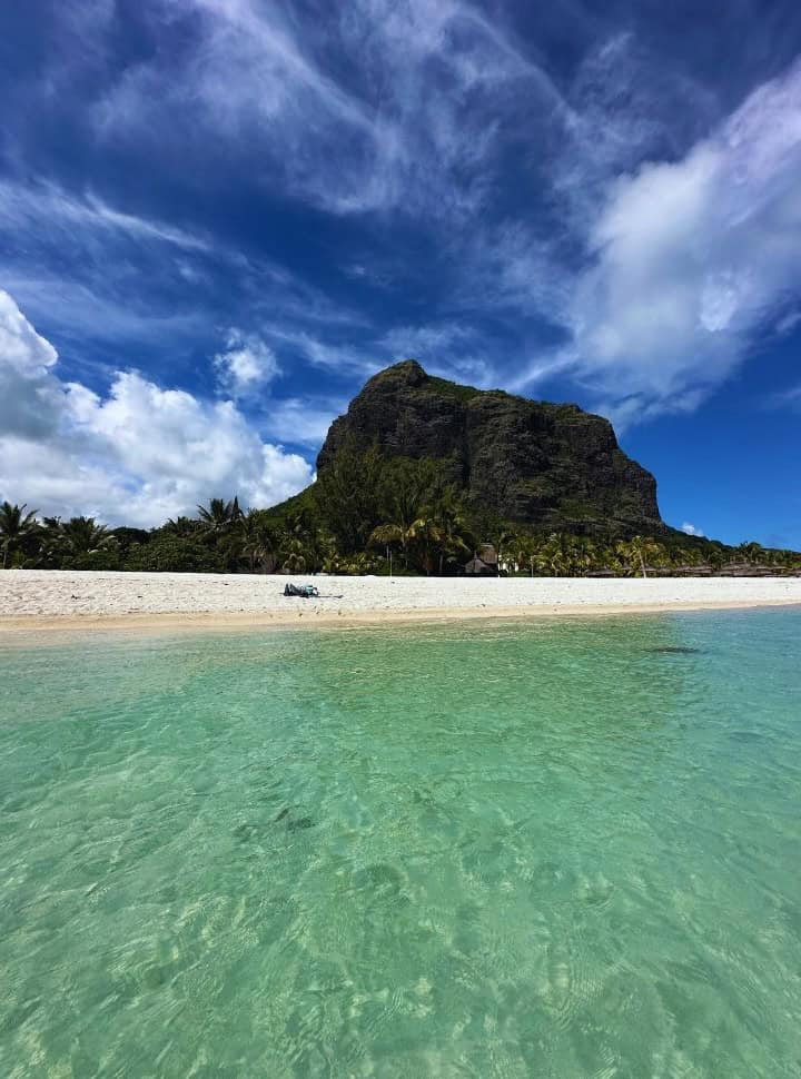large rock mountain set behind the tropical beach with rows of palm trees, white sand and clear turquoise ocean water In Mauritius