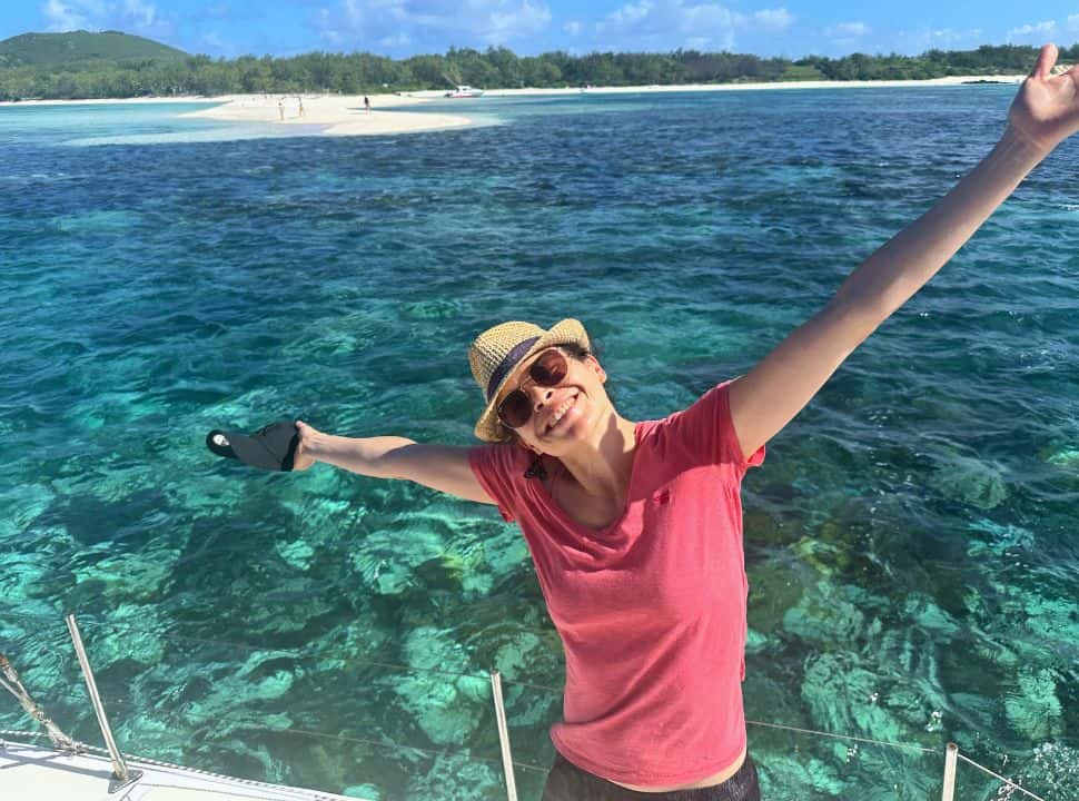 woman posing with arms in the air on a boat sailing on turquoise blue water and in the back a bright white sand bank set on a small island in Mauritius