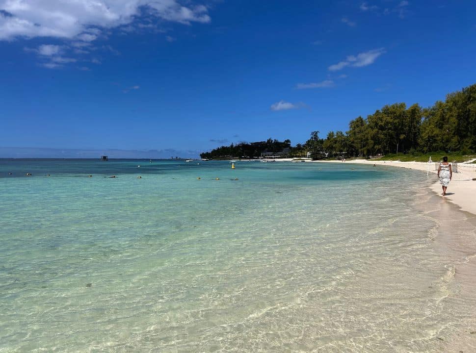 tropical beach with clear turquoise water and a white sand, a woman is walking along the shore with lush vegetation on her right at Belle Mare Mauritius