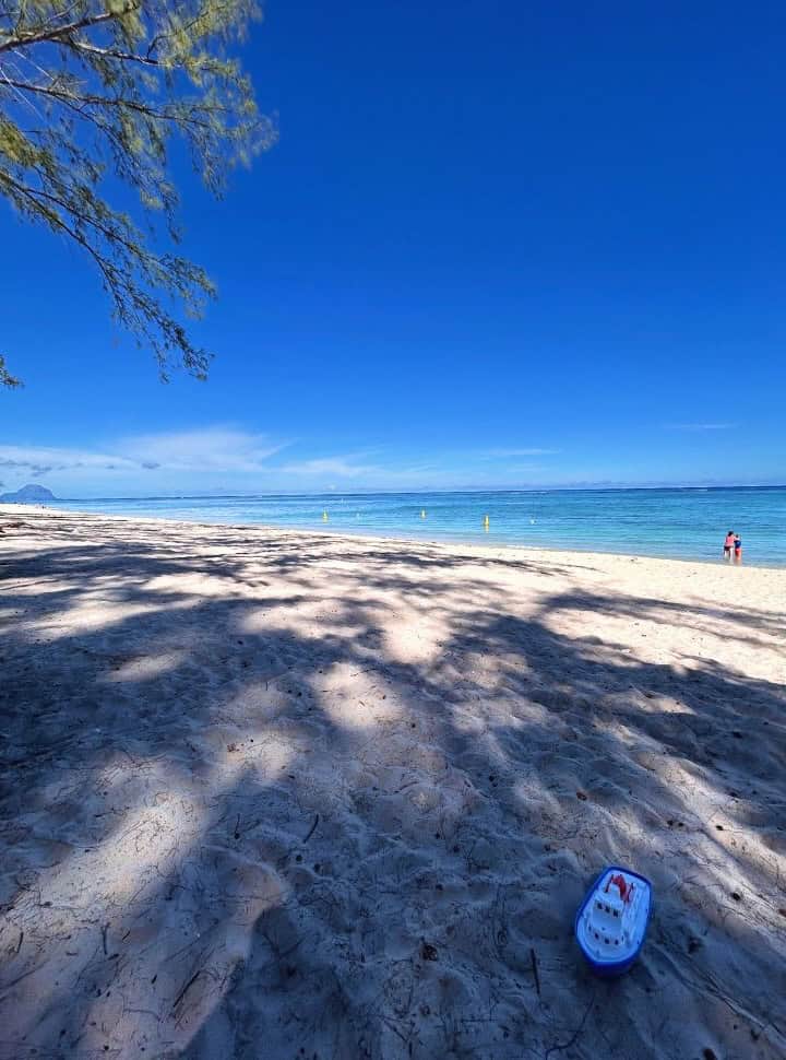mother and son standing in the blue water at a soft sanded beach, a toy boat is left behind in the shade at Flic en Flac Mauritius