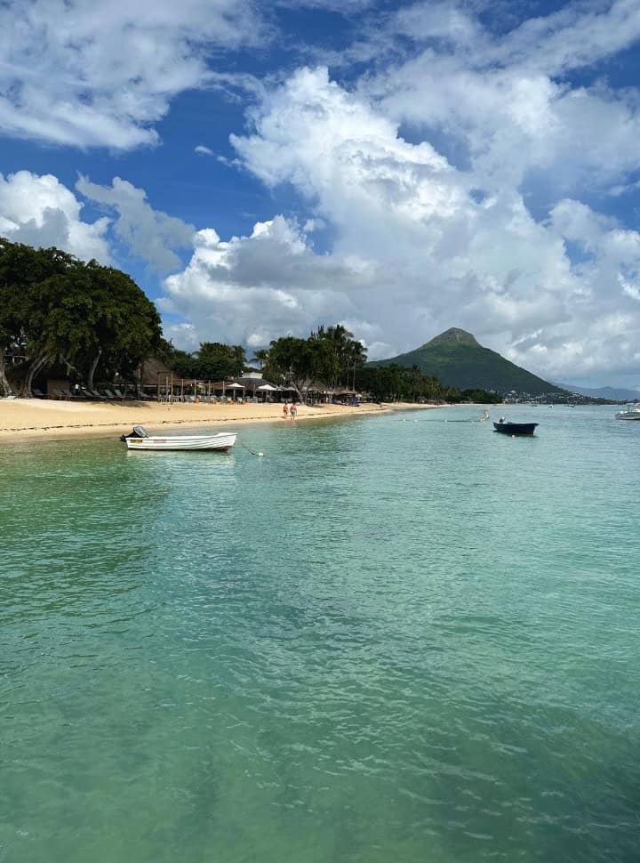 boats anchored along the coast of a tropical beach nearby Flic en Flac, a couple is walking along the shore passing sun loungers. In the back you can see the peak of a mountain