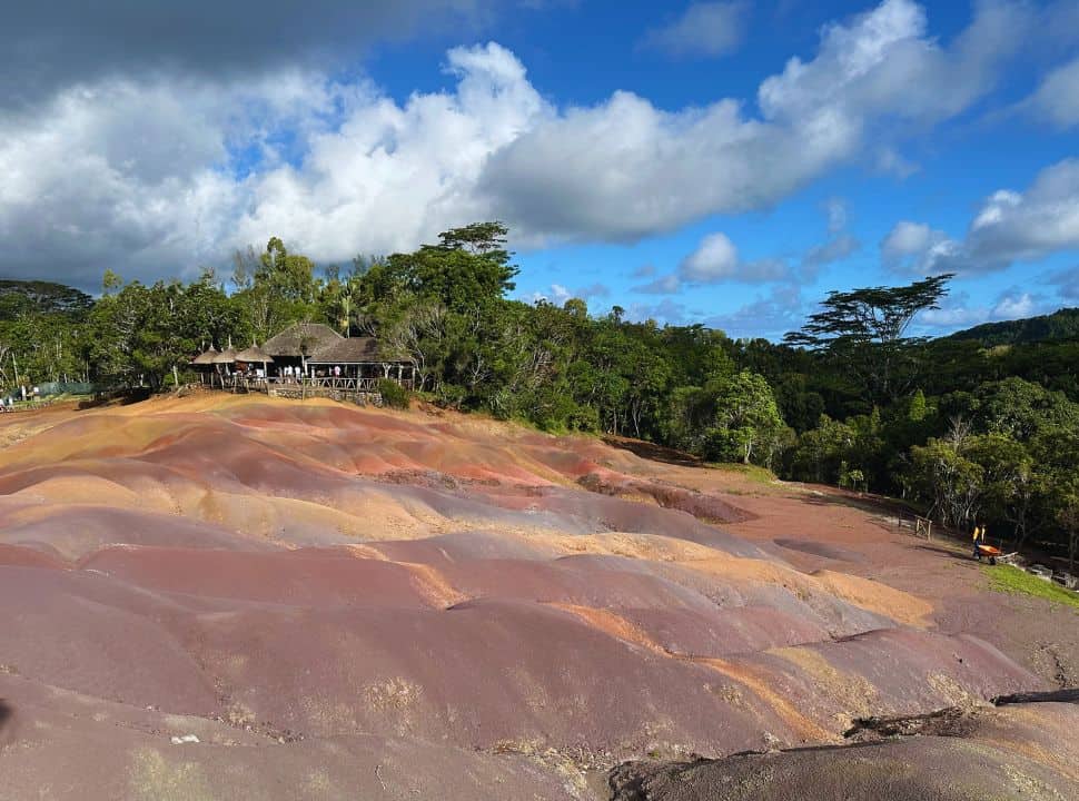 rocky hill with all shades of red and yellow surrounded by green trees, there is a viewing point in a distance at the Chamarel Seven Colored Earth Geopakr