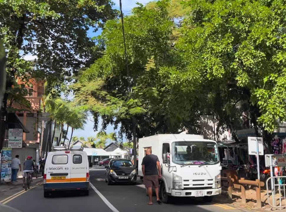 street in Grand Baie Mauritius with lorries and cars, shops selling dresses and a tour company all set under large green trees