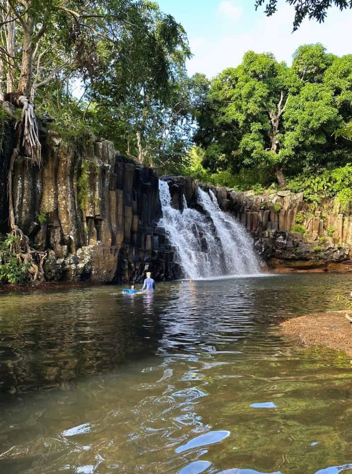 man and boy swimming in the waterfall pool, in the back is a beautiful waterfall surrounded by large trees in the south of Mauritius
