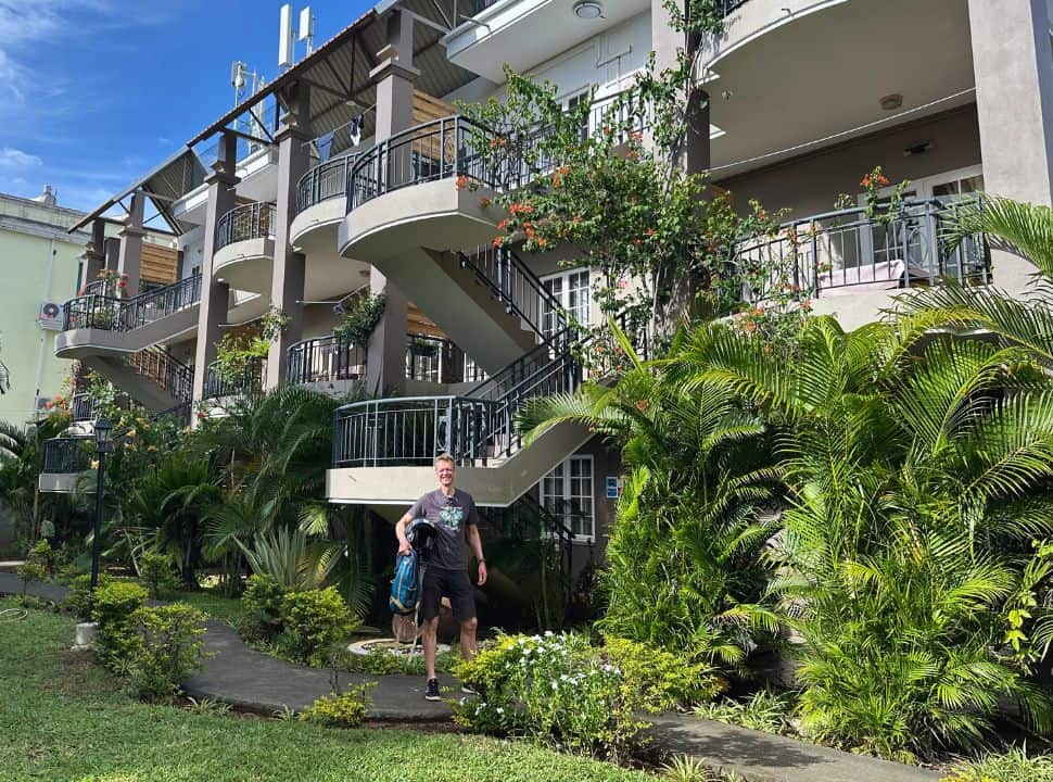 man standing in front of a two story accommodation with balconies and decorated with green tropical plants in pereybere Mauritius