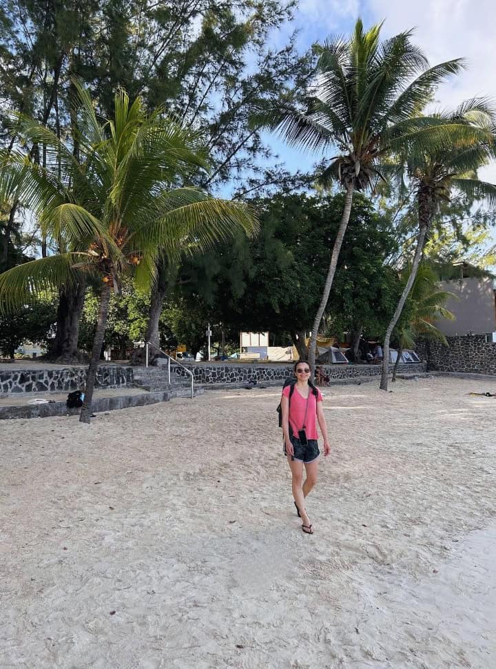 woman walking on quiet Pereybere beach, palm trees and a park like area in the background