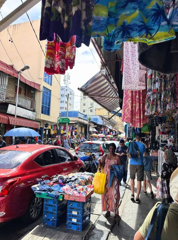 people walking on the side walk where you can also find goods for sale, clothing next to parked cars and displayed high above the pavement where people walk under it in Port Louis Mauritius