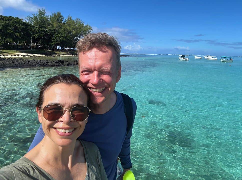 couple posing for a selfie with in the background clear ocean water in the color turquoise, boats are anchored near the shore in Mauritius