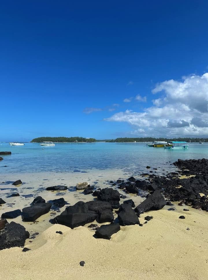 view of the water from a beach with volcanic rocks, white sand and clear water. Rocks are visible in the water in the distance. Boats are anchored in the water with two islands in the back