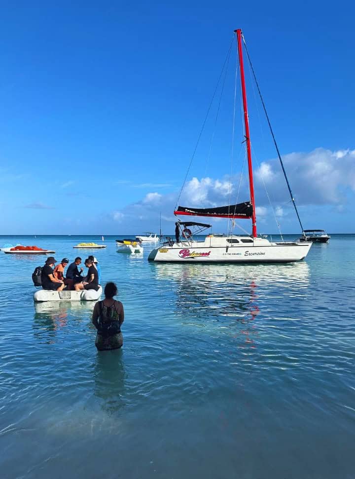 woman standing in the shallow ocean water watching a small boat with passengers heading towards a catamaran anchored just offshore of Pereybere beach Mauritius