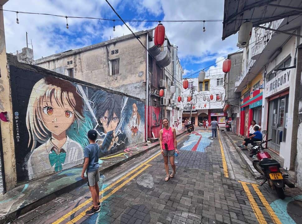 woman walking through an alley with japanese manga murals, chinese lanterns hanging over the street and old houses in Port Louis Chinatown