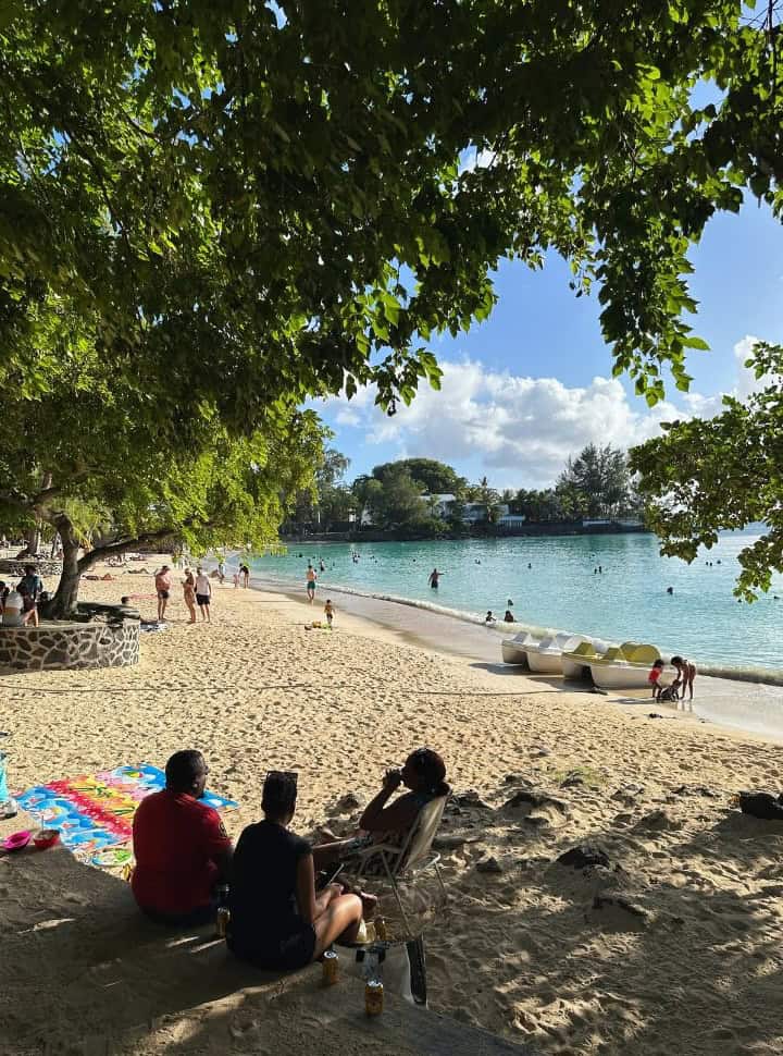 a beach with people sitting in the shade of the tree, paddle boats along the shore, swimmers in the water along the far edge there is accommodation, Grand Baie Mauritius looks less laid back