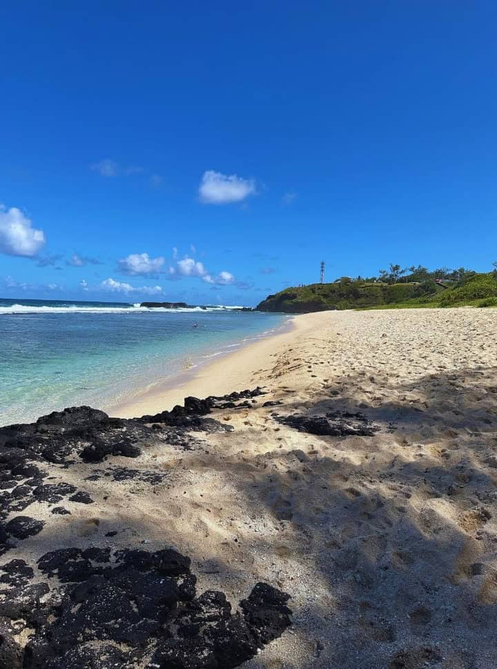 deserted white soft sanded beach in Mauritius along the blue water, in the distance is a headland covered with vegetation, there are black rocks visible in the sand to 
