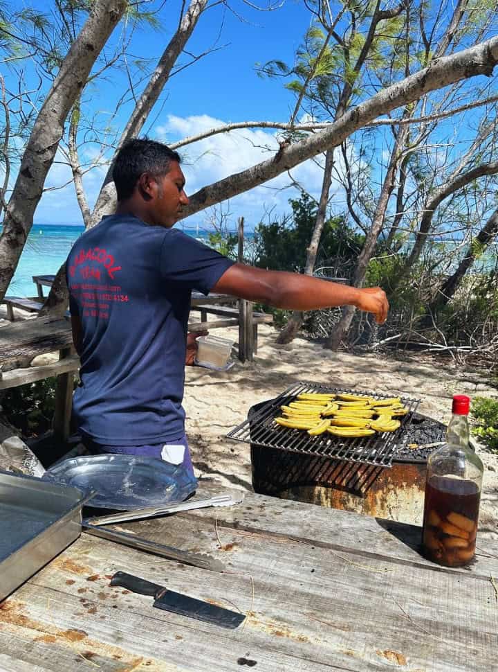man standing at a barbecue with plantains, adding sugar while a large bottle of rum is standing nearby. A tour company in Mauritius 