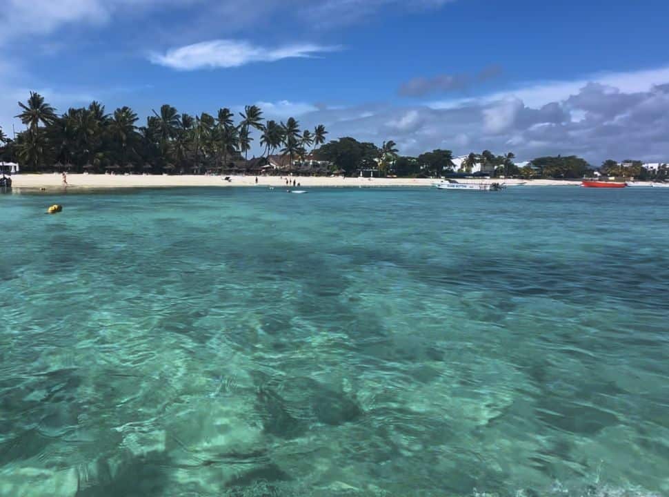 view of a long stretched white beach with palm trees and accommodation,  picture taken from the water which is crystal clear and in all sorts of turquoise colours located north west of Mauritius