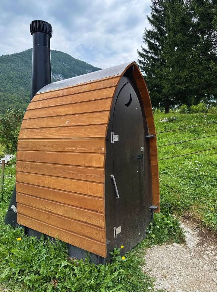 public toilet that looks like a small cabin, with wooden walls and a chimney. Door is decorated with a moon. It's an eco toilet set in the field in Slovenia
