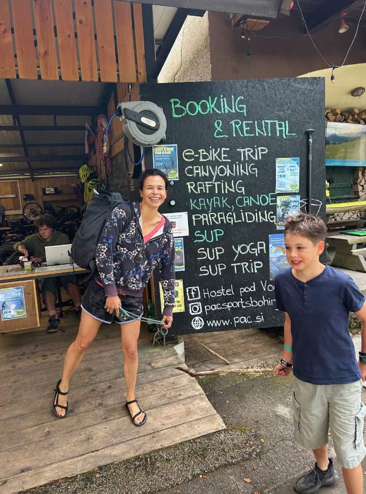 mother and son standing in front of a rental company at lake bohinj, signs showing all the things you can do from paragliding, SUP trip and SUP yoga