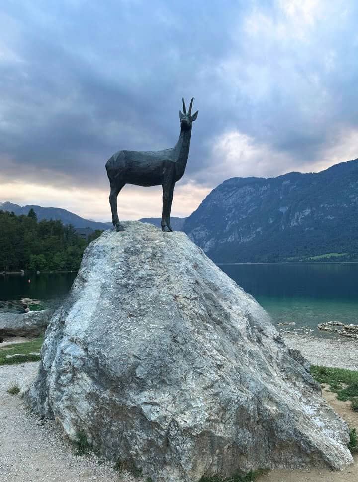 a statue of a deer set on a tall rock right on the shore of lake bohinj in Slovenia