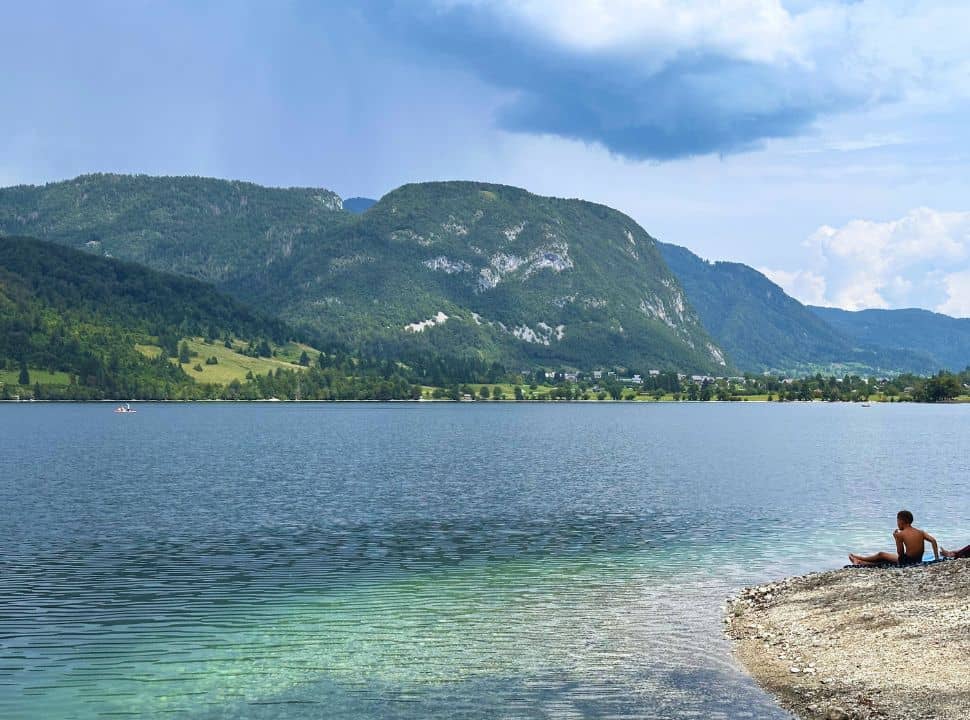 boy sitting on a pebbled beach along a lake with stunning colored water, in the back you can see the mountain slopes surrounding lake Bohinj Slovenia