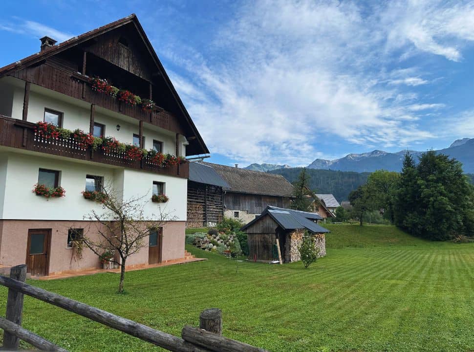 traditional Slovenian house set along a green grass, shed is found nearby and it is surrounded by mountains, nearby Lake Bohinj