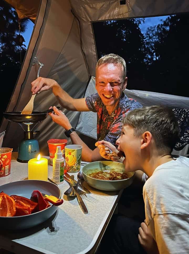 father cooking something on a camping stove in a tent while son is enjoying his dinner