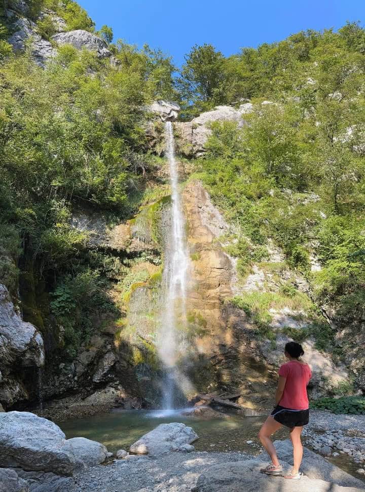 woman looking at a waterfall with a small pool surrounded by green vegetation in Slovenia