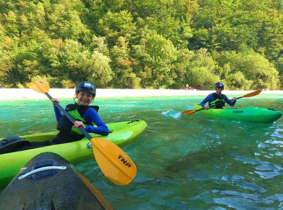 father and son in a kayak paddling in the Soca River with a white river beach in the back and green vegetation