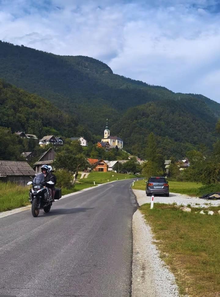 motorcycle driving along the mai road, in the background a traditional Slovenian church is visible perched on a hill top. 