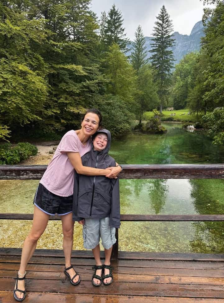 mother and son posing for a picture at a bridge that crosses a green colored yet crystal clear river surrounded by pine trees at Lake Bohinj Slovenia