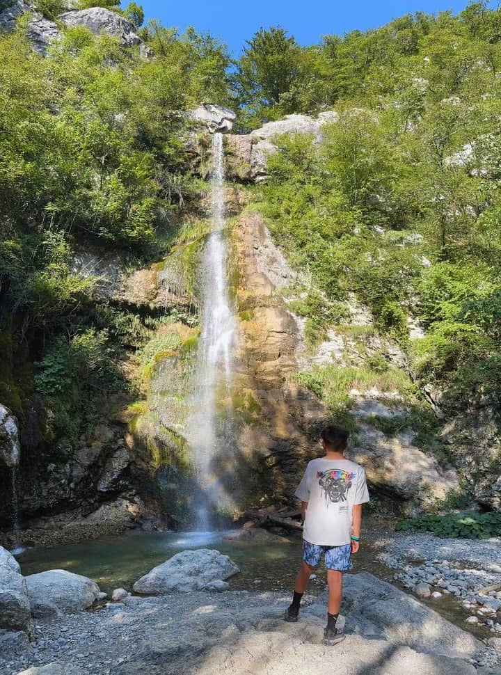 boy standing on a bolder while looking at the waterfall and the pool, the rocks around the waterfall are lush green, Slovenia waterfall Beri