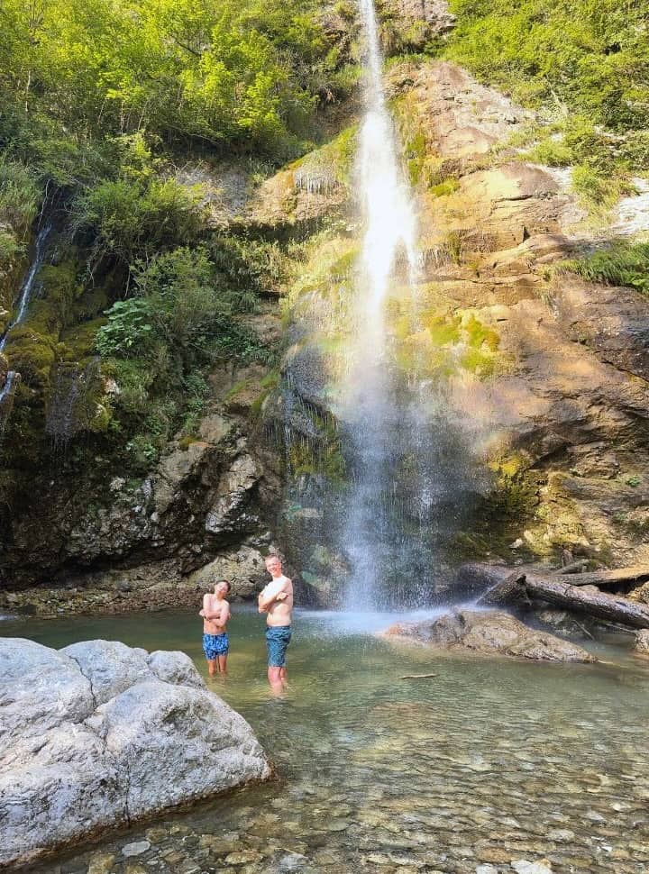 father and son posing for a picture while standing in the waterfall pool, a tall waterfall in the back near Tolmin Village Slovenia