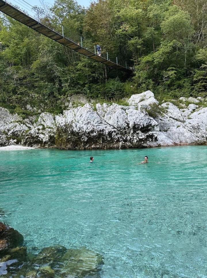 mother and son swimming in the stunning blue water of Soca River, white rocks on the edge and a wooden bridge above, nearby Kozjak Waterfall Slovenia
