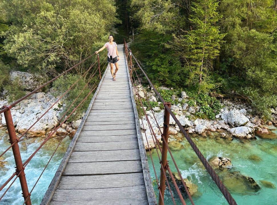 woman standing on a wooden bridge in Soča Valley Slovenia hat crosses a river with crystal clear emerald water, in the back pine trees