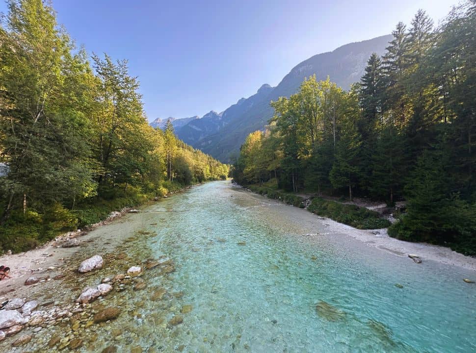 view of a clear blue river flowing passed lush green trees on the river banks, white stones are scattered along the river and in the distance there is a mountain range at Soča Gorge Slovenia
