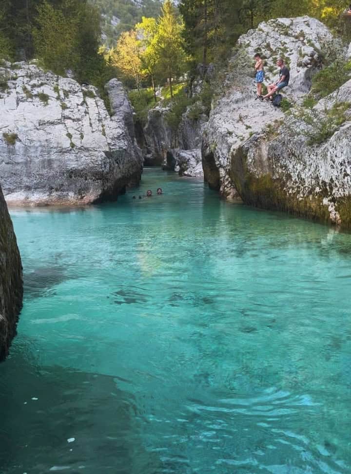 river flowing within a the Soca Gorge, there are people swimming and standing along the edge on the tall white rocks, the water is clear and looks unreal. 