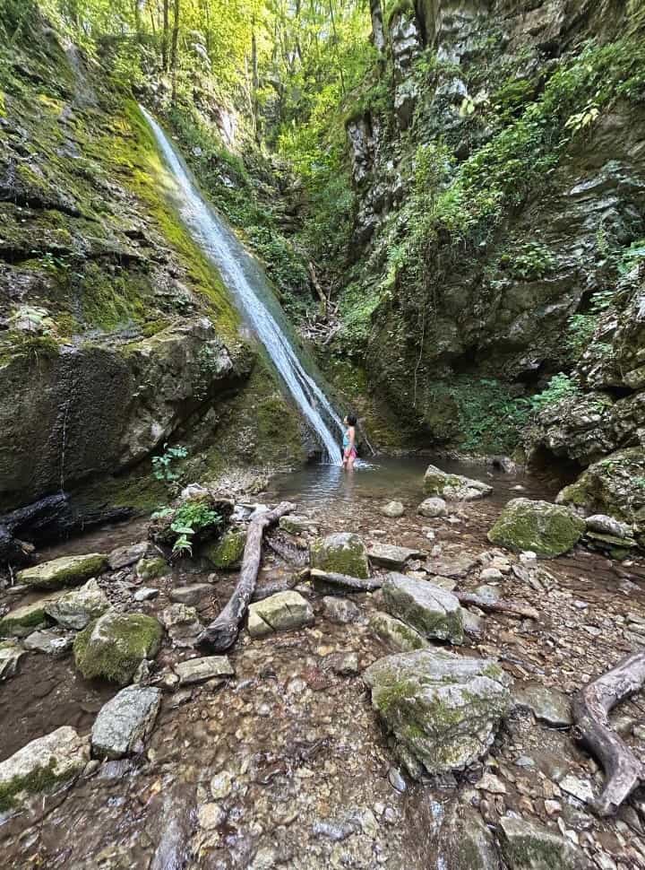 woman looking up to a waterfall while standing in the bottom in a waterfall pool surrounded by rocks and lush forest near Kobarid Slovenia