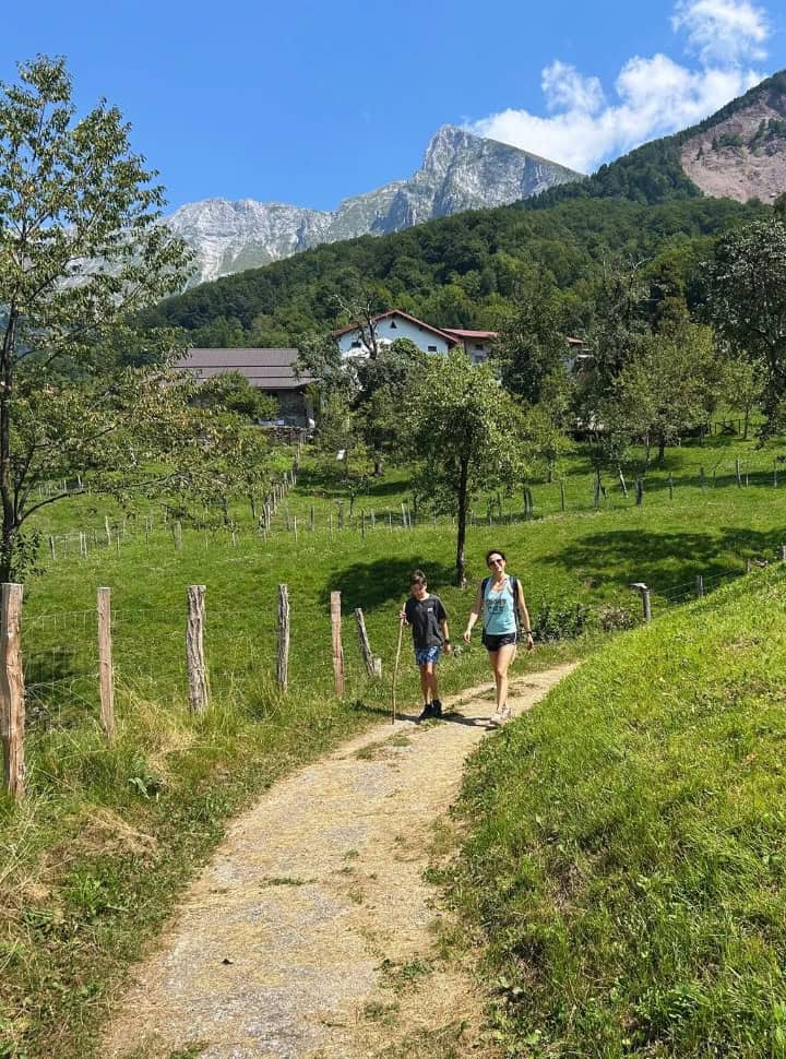 mother and son walking along a path near a farm with apple trees in the field and mountain tops visible in the back in Slovenia