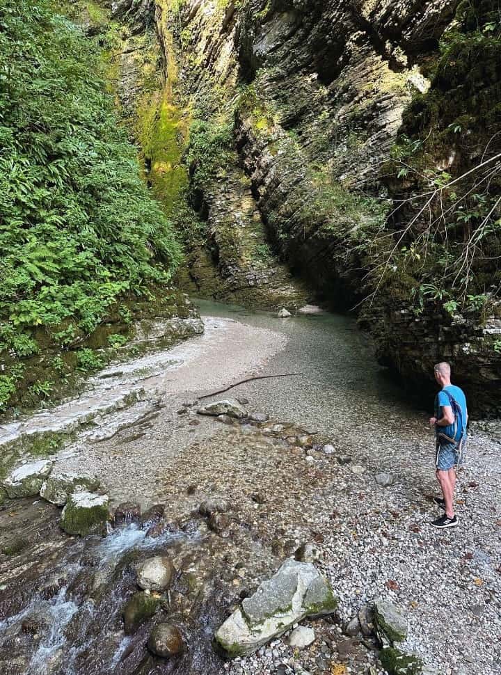 man standing near the clear river within a gorge that is overgrown with moss, plants and trees just before arriving at Kozjak Waterfall Slovenia