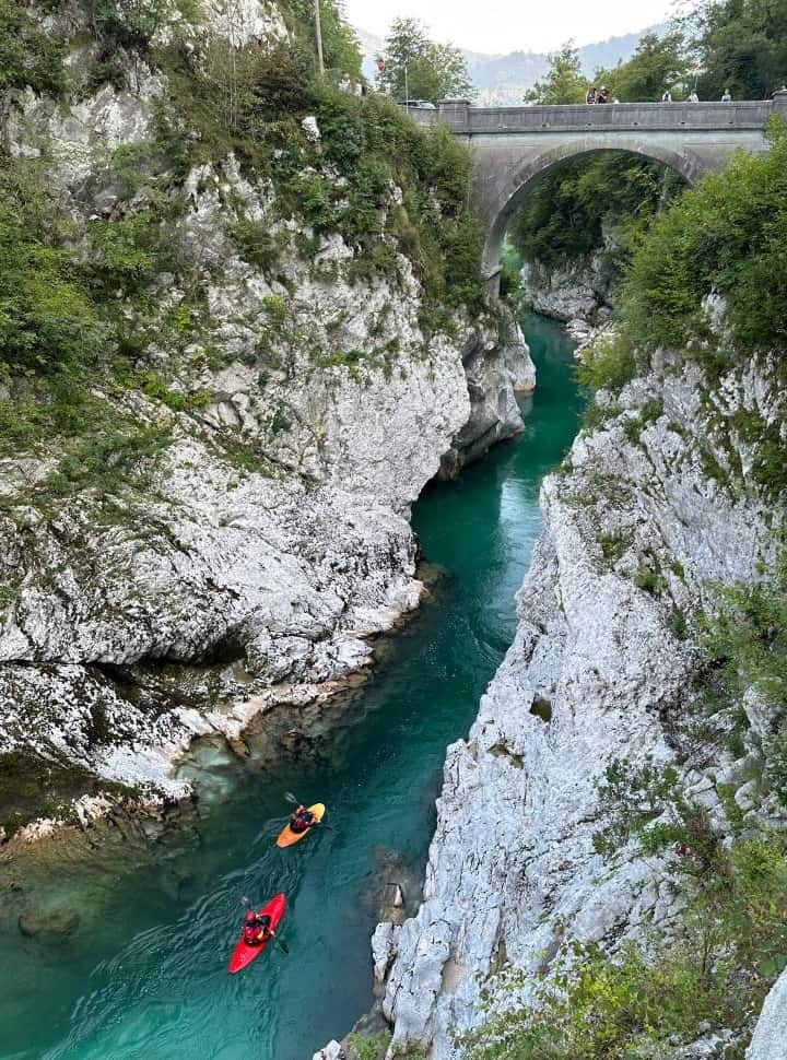 view of the napoleon bridge above the emerald green river set within the white stone gorge, people are crossing the bridge while kayakers are approaching it on the river near Kobarid Slovenia
