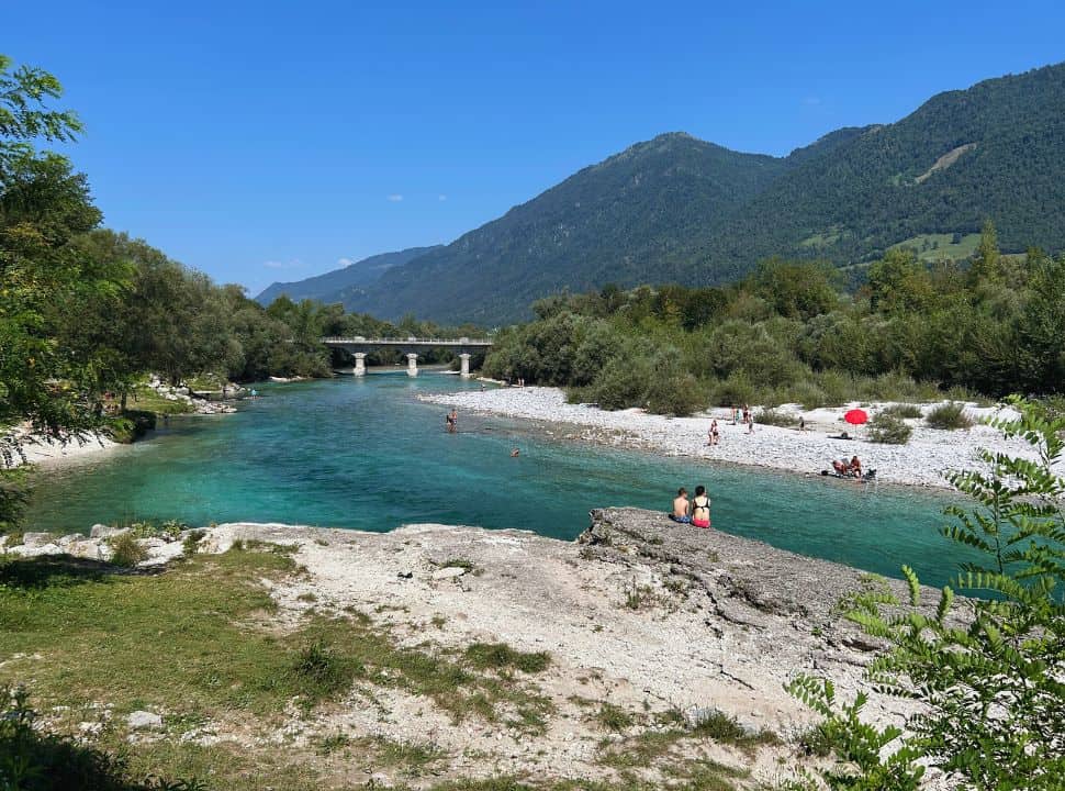 view of a pebbled beach along soca river on one side and rocky service on the other, people are swimming in the crystal clear blue river near Tolmin village Slovenia