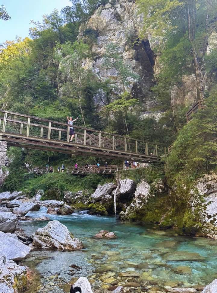 man crossing one of the many bridges set within the Tolmin Gorge, the setting is beautiful with green vegetation along the gorge walls and a clear blue river running through. 