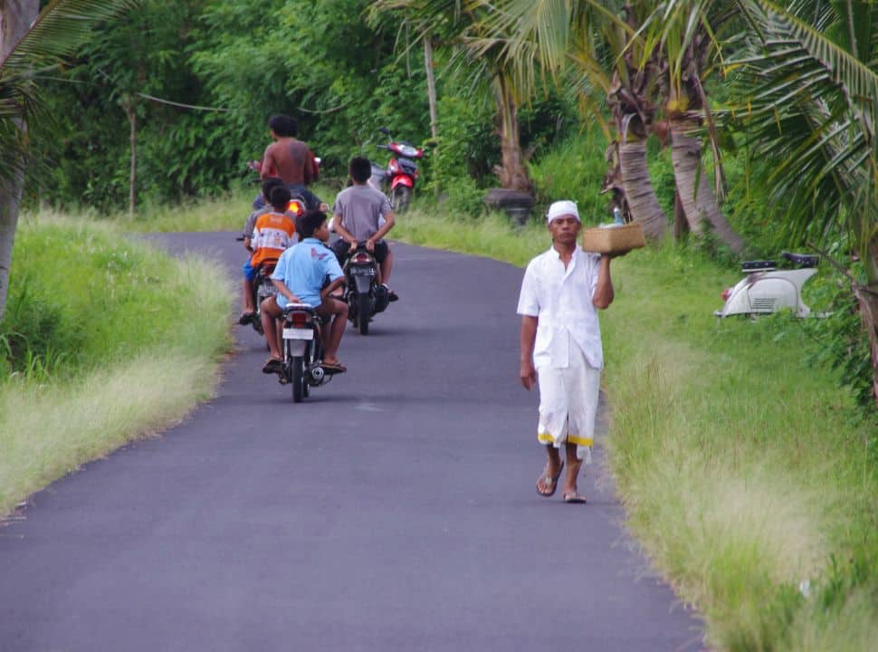 a priest walking along the side of the road holding a basket with items for prayers, a group of young kids are passing him on a motor scooter in a quiet street in Sidemen Bali