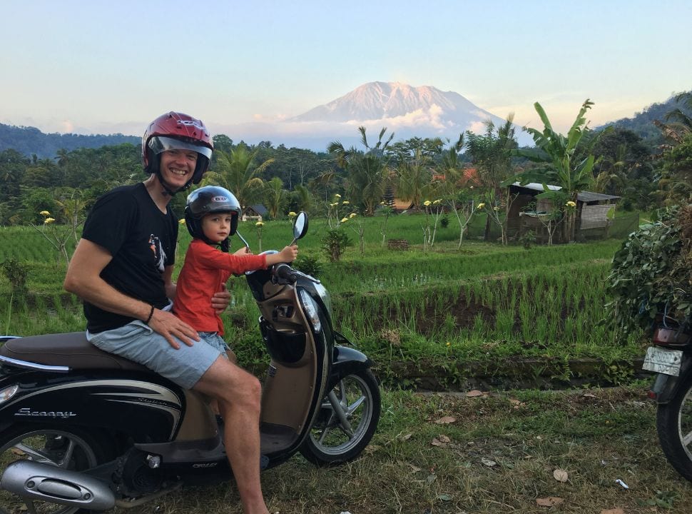 father and son posing on a motor scooter with the peak of mount agung basking in the sun while the valley is green with rice fields and trees. 